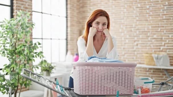 Young redhead woman leaning on basket with clothes looking upset at laundry room - Footage, Video