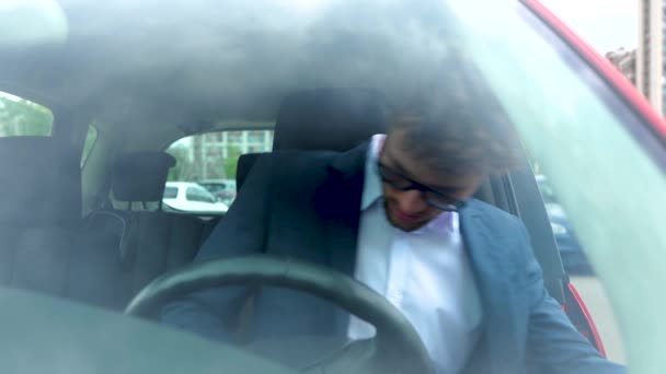 Stílusos fiatalember szemüvegben, kinyitja az ajtót és beszáll a kocsiba, biztonsági övvel. Személyek védelme járművekben. Közlekedés, üzlet, biztonság és emberek koncepciója. Lassú mozgás. - Felvétel, videó