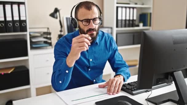 Fiatal spanyol férfi üzletember számítógéppel és fejhallgatóval jegyzetel az irodában - Felvétel, videó