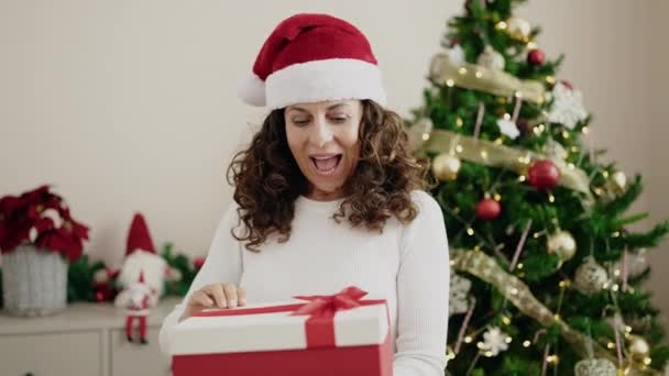 Hispanische Frau mittleren Alters packt Geschenk aus, das zu Hause am Weihnachtsbaum steht - Filmmaterial, Video