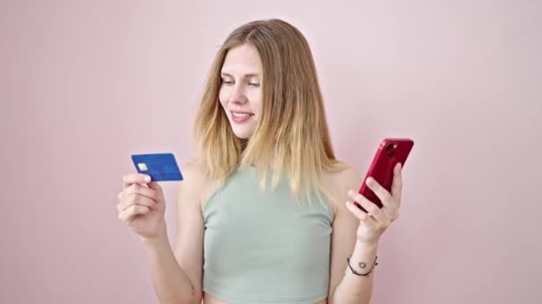 Молодая блондинка держит смартфон и кредитную карту с жестом победителя на изолированном розовом фоне - Кадры, видео