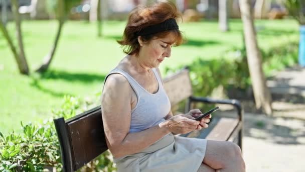 Donna di mezza età utilizzando smartphone con espressione grave seduto sulla panchina al parco - Filmati, video