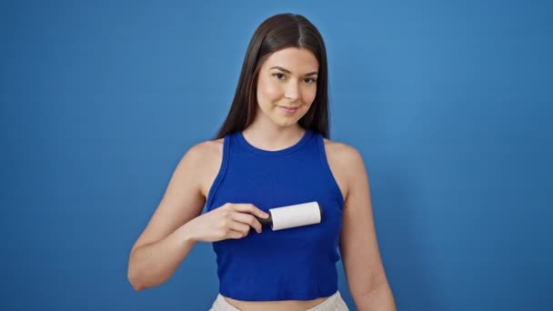 Junge schöne hispanische Frau lächelt selbstbewusst Reinigungs-T-Shirt mit Tierhaarrolle über isolierten blauen Hintergrund - Filmmaterial, Video