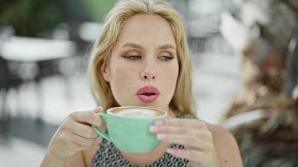 Nuori blondi nainen puhaltaa ilmaa kuppi kahvia hymyillen kahvilan terassilla - Materiaali, video