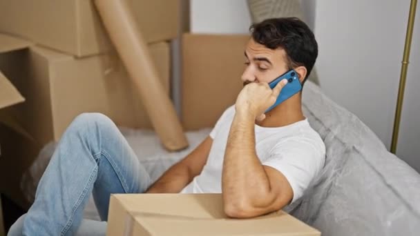 Hombre hispano joven hablando en un smartphone sentado en un sofá en un nuevo hogar - Metraje, vídeo