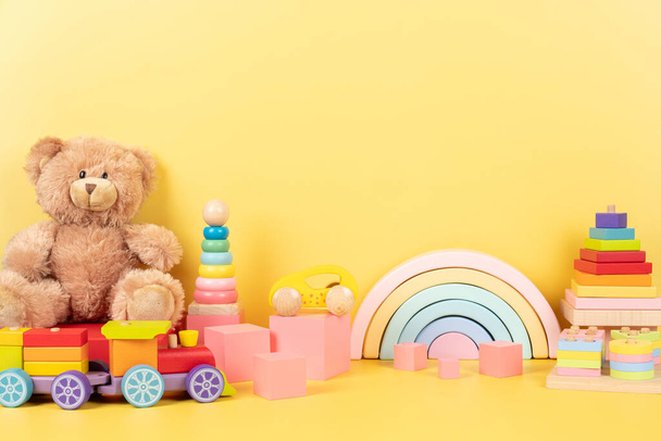 Collection de jouets éducatifs pour enfants. Ours en peluche, arc-en-ciel en bois, train, empilant des cubes roses pyramidaux sur fond jaune. Vue de face. - Photo, image