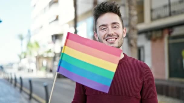Jonge Spaanse man glimlacht vol vertrouwen met regenboogvlag op straat - Video