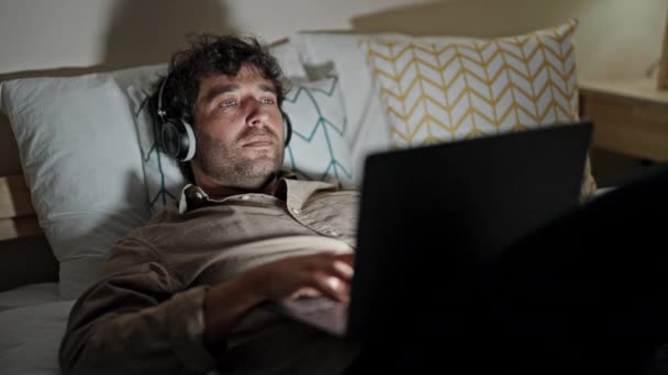 Hombre hispano joven usando portátil y auriculares relajados en la cama en el dormitorio - Imágenes, Vídeo