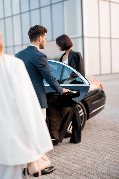 Businessman and businesswoman walk together to car on parking lot near modern building, going to drive by luxury car. Man opens vehicle door - Фото, изображение