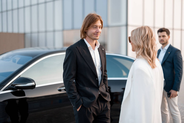 Eleganccy biznesmeni rozmawiają w pobliżu luksusowego samochodu na zewnątrz. Partnerzy biznesowi rozmawiają po wieczornej imprezie na parkingu - Zdjęcie, obraz