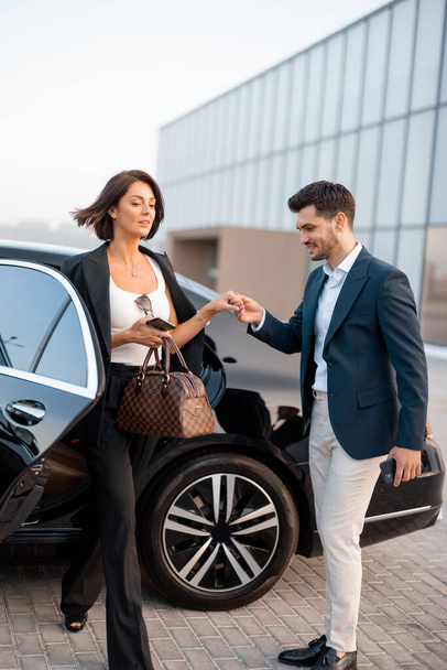 Businessman helps gently a woman to get out of a car, arrived by luxury vehicle for some event at evening. Concept of transportation and business lifestyle - Zdjęcie, obraz