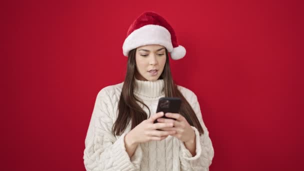 Mujer hispana hermosa joven usando sombrero de Navidad usando teléfono inteligente sobre fondo rojo aislado - Imágenes, Vídeo