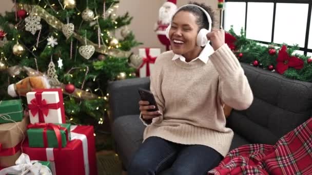 African american woman dancing and listening to music sitting on sofa by christmas tree at home - Footage, Video
