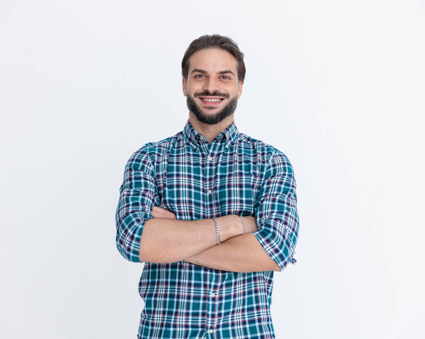 portret van knappe jongeman met baard in geruite overhemd die armen kruist en glimlacht voor grijze achtergrond in studio - Foto, afbeelding