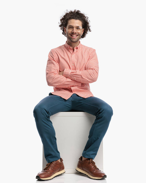 excited young man with curly hair and glasses crossing arms, sitting and laughing in front of white background in studio - Foto, Bild