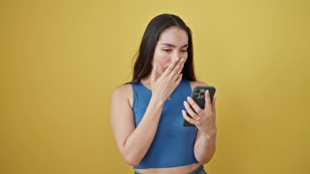 Junge schöne hispanische Frau, die ihr Smartphone benutzt und Nein sagt, überrascht mit dem Kopf über den isolierten gelben Hintergrund - Filmmaterial, Video