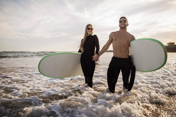 Xtreme Sports (s. Nuori urheilullinen pari surffaa yhdessä rannalla, hymyilevän miehen ja naisen muotokuva märkäpuvut kädessä ja surffilaudat kädessä, tuhatvuotiset surffaajat seisomassa vedessä - Valokuva, kuva