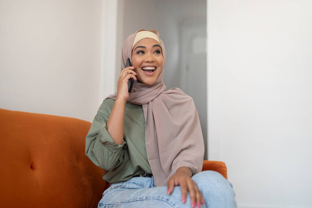Matkaviestintä. Iloinen musta nainen hijabissa puhuu matkapuhelimessa istuu sohvalla kotona, nauttii keskustelusta, kommunikoi ystävien kanssa älypuhelimen kautta, katsomalla sivuun - Valokuva, kuva