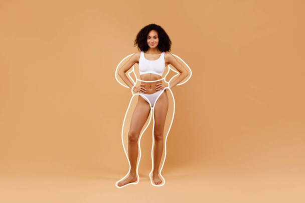Body sculpting. Young sporty black woman in underwear with drawn outlines around figure stand isolated over beige background. Beautiful lady demonstrate result of weightloss, collage with copy space - Photo, Image