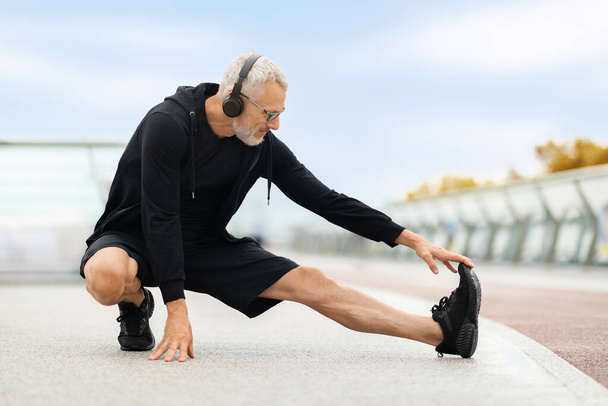 Sport, zdravý životní styl. Odhodlaný dobře padnoucí starší muž v černém sportovním oblečení pomocí bezdrátových sluchátek natahujících tělo nohy před jogging po mostě na podzim, mají venkovní cvičení, kopírovací prostor - Fotografie, Obrázek