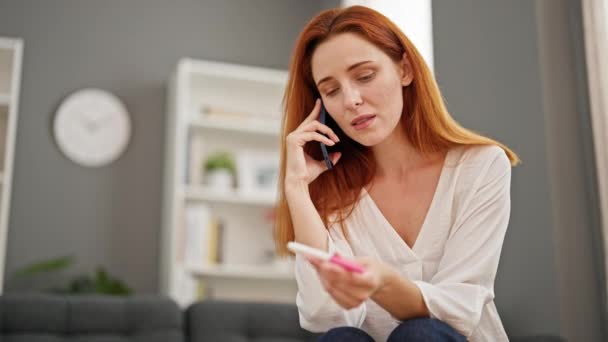 Young redhead woman talking on smartphone holding pregnancy test looking upset at home - Footage, Video