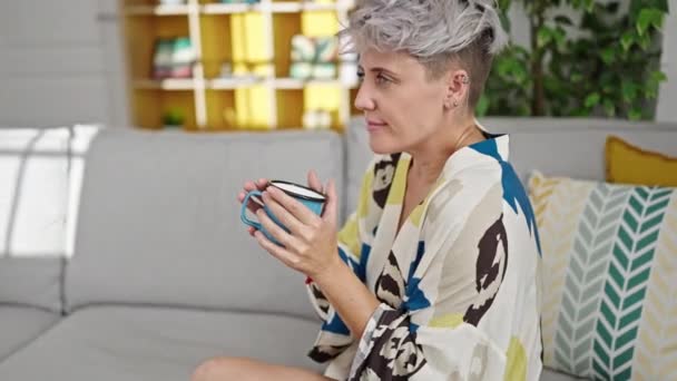 Nuori nainen pitämällä kuppi kahvia istuu sohvalla kotona - Materiaali, video