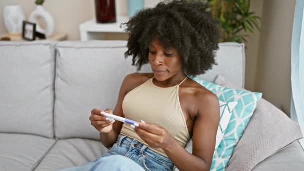 Mujer afroamericana sonriendo confiada sosteniendo prueba de embarazo sorprendida en casa - Imágenes, Vídeo