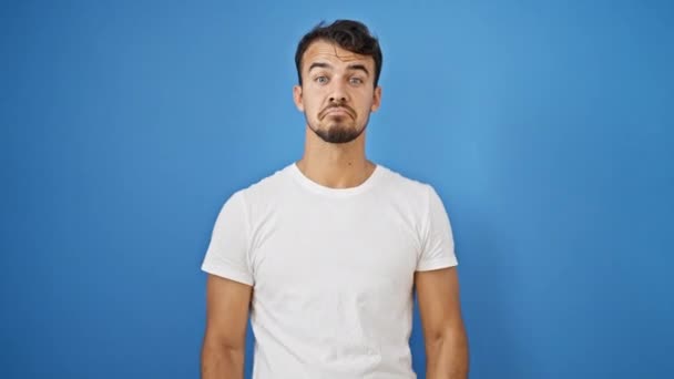 jonge latino man staan onwetend over geïsoleerde blauwe achtergrond - Video