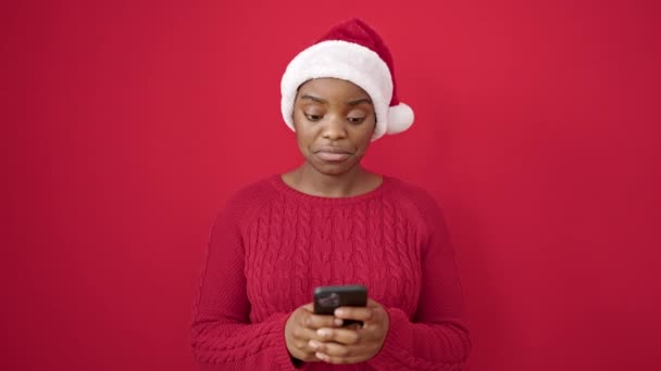Afroamerikanerin mit Weihnachtsmütze zeigt auf Smartphone vor isoliertem roten Hintergrund - Filmmaterial, Video
