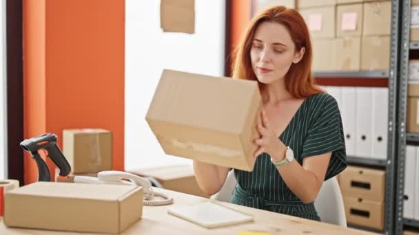 Young redhead woman ecommerce business worker writing on notebook holding package at office - Footage, Video