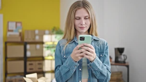 Junge blonde E-Commerce-Kauffrau lächelt mit Smartphone im Büro - Filmmaterial, Video