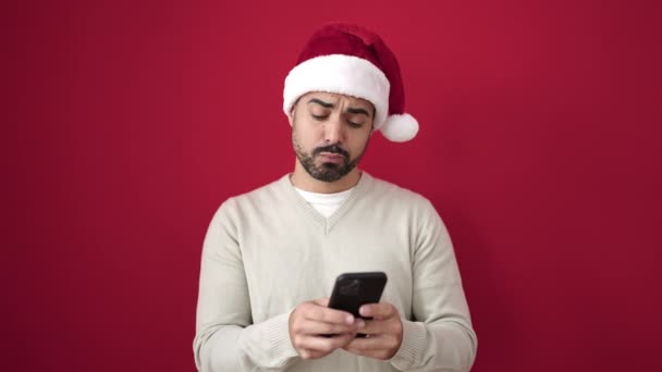 孤立した赤い背景の上にスマートフォンを使用してクリスマスの帽子をかぶっている若いヒスパニック男性 - 映像、動画