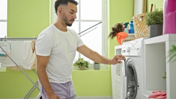 Молодий арабський чоловік миє одяг за допомогою смартфона в пральні - Кадри, відео
