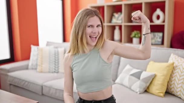 Junge blonde Frau lächelt selbstbewusst und macht starke Geste mit Arm zu Hause - Filmmaterial, Video