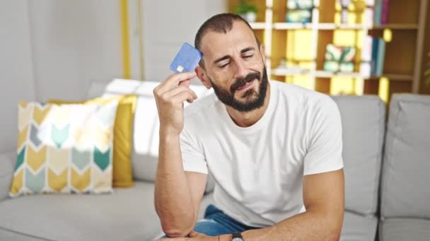 Joven hispano sosteniendo tarjeta de crédito sentado en sofá pensando en casa - Imágenes, Vídeo