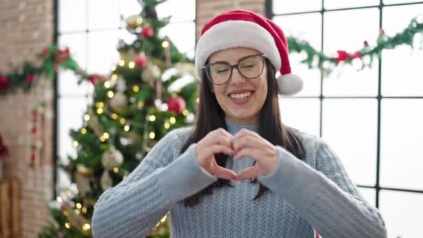 Joven mujer hispana haciendo gesto de corazón de pie junto al árbol de Navidad en casa - Metraje, vídeo