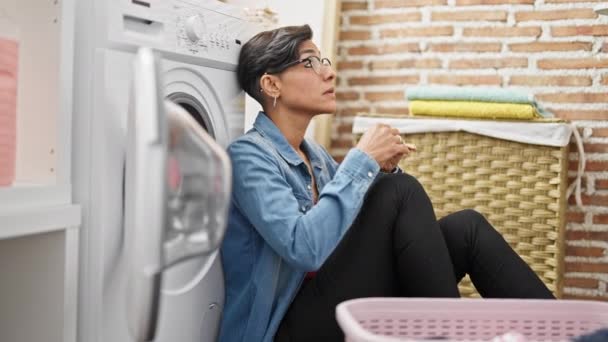 Молода красива іспаномовна жінка спирається на пральну машину з серйозним виразом в пральні - Кадри, відео