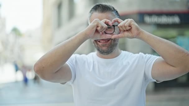 Hombre de pelo gris sonriendo confiado haciendo gesto de corazón con las manos en la calle - Imágenes, Vídeo