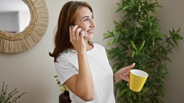 Mujer joven sosteniendo taza de café hablando en el teléfono inteligente en casa - Imágenes, Vídeo