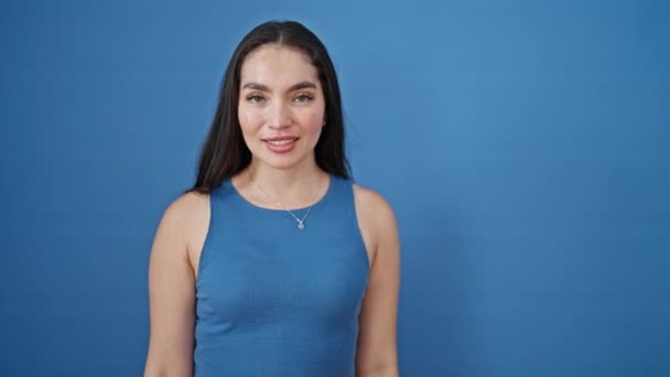 Νεαρή όμορφη ισπανόφωνη γυναίκα χαμογελά με αυτοπεποίθηση κάνοντας στρατιωτικό χαιρετισμό πάνω από απομονωμένο μπλε φόντο - Πλάνα, βίντεο