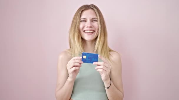 Молодая блондинка улыбается уверенно указывая на кредитку на изолированном розовом фоне - Кадры, видео