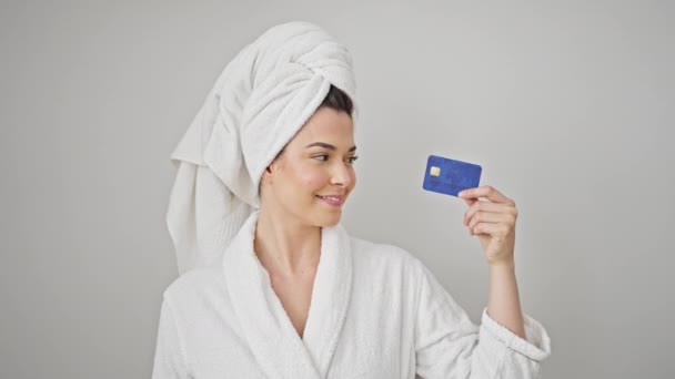 Fiatal gyönyörű spanyol nő visel fürdőköpeny gazdaság hitelkártya mosolyogva elszigetelt fehér háttér - Felvétel, videó