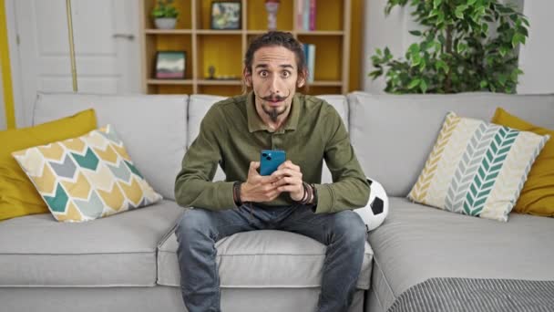若いですヒスパニック男サポートサッカーチーム座っています上のソファに家 - 映像、動画
