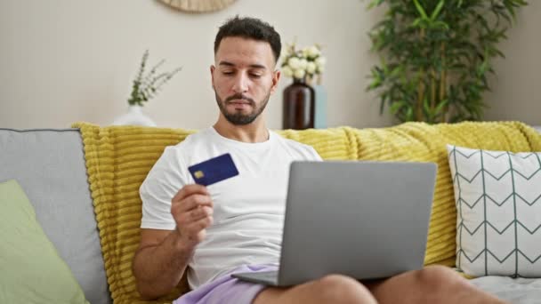 Jovem árabe homem compras com laptop e cartão de crédito sentado no sofá em casa - Filmagem, Vídeo