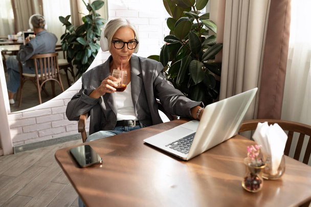 uroczy stylowy dojrzały biznes kobieta w okularach ubrany w szarą kurtkę spędza czas w kawiarni siedzi z laptopem. - Zdjęcie, obraz