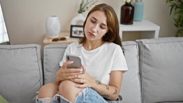 Mujer joven usando teléfono inteligente sentado en el sofá buscando molesto en casa - Metraje, vídeo
