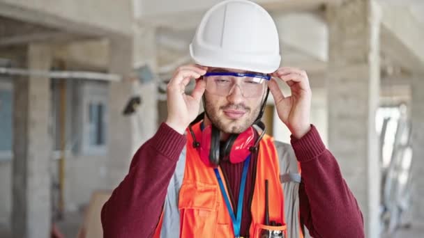 Jonge Spaanse man bouwer glimlacht vol vertrouwen het nemen van een bril af op de bouwplaats - Video