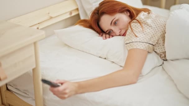 Νεαρή κοκκινομάλλα γυναίκα που βλέπει τηλεόραση ξαπλωμένη στον καναπέ κουρασμένη στο υπνοδωμάτιο - Πλάνα, βίντεο