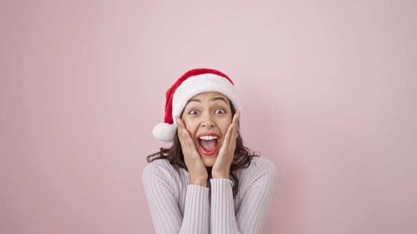 Mujer hispana hermosa joven expresión sorpresa usando sombrero de Navidad sobre fondo rosa aislado - Imágenes, Vídeo