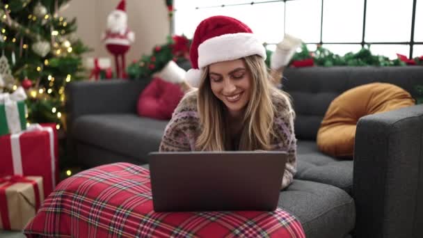 Νεαρή ξανθιά γυναίκα που χρησιμοποιεί φορητό υπολογιστή ξαπλωμένη στον καναπέ δίπλα στο χριστουγεννιάτικο δέντρο στο σπίτι - Πλάνα, βίντεο
